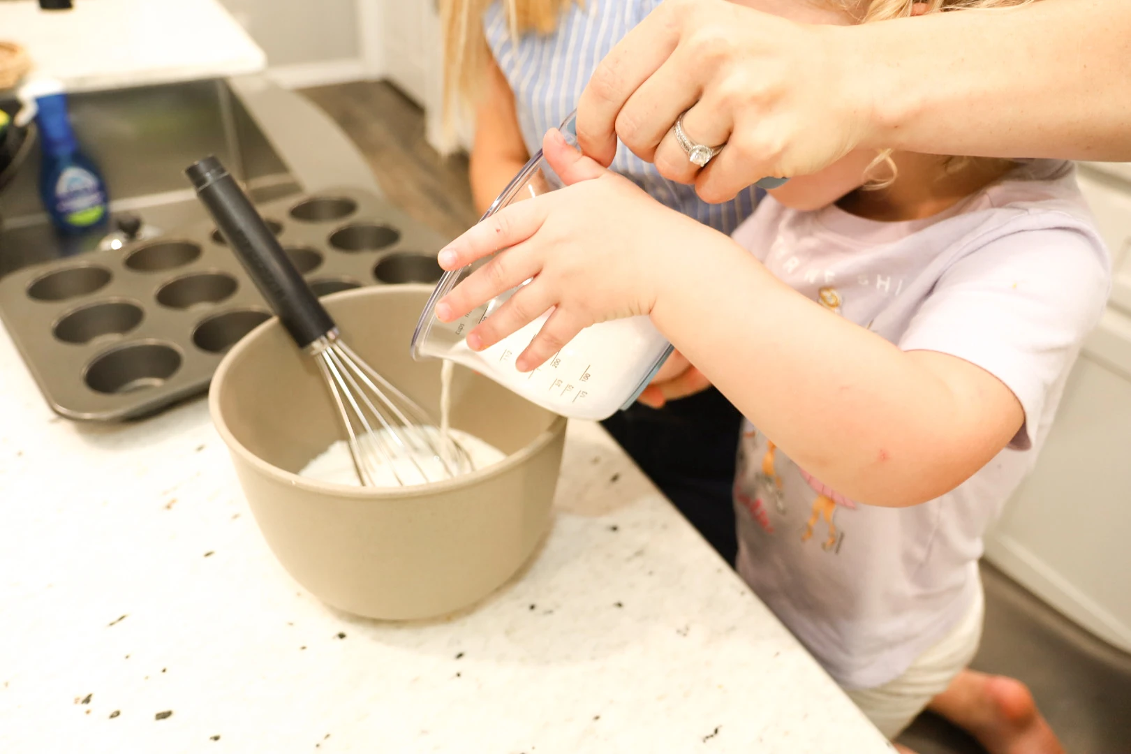 A mother and young child smiling while pouring batter into a bowl together.