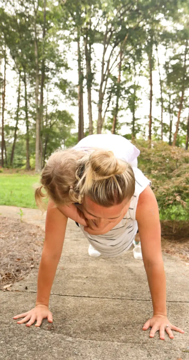 A mom doing push-ups on a trail in the woods while a toddler hangs onto her back.